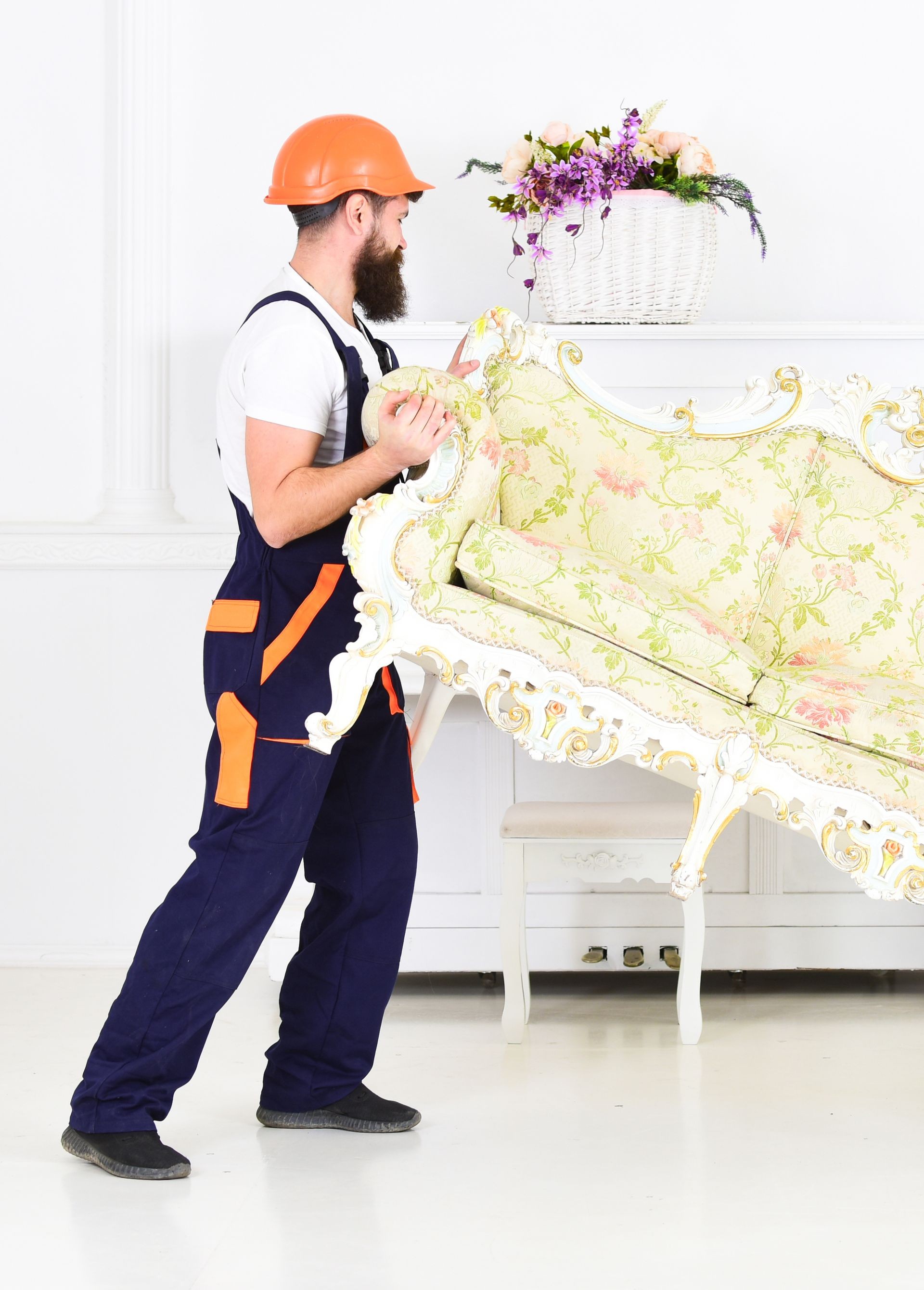 Loader moves sofa, couch. Man with beard, worker in overalls and helmet lifts up sofa, white background. Courier delivers furniture in case of move out, relocation. Delivery service concept.